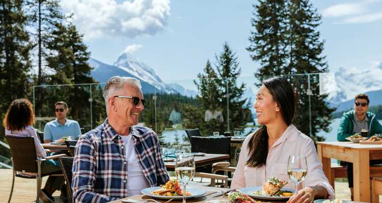 Groups of people sit at patios tables outside of a building, mountains in the background.