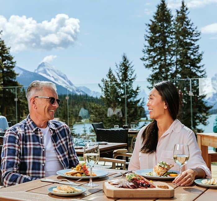 Couple sits on The View patio with fish on their plates and Maligne Lake behind them
