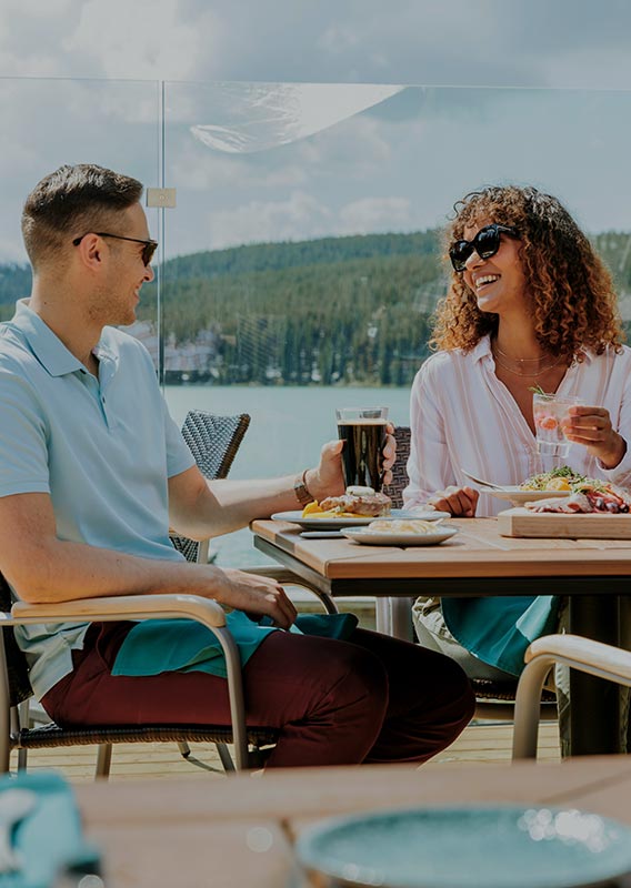 Two people sit at a dining table on a patio overlooking a blue lake surrounded by forested mountains