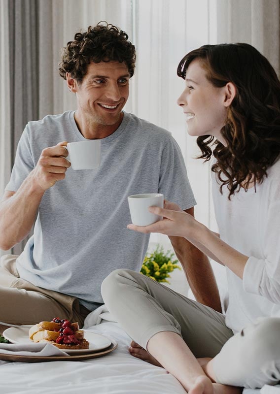 Two people sit on a hotel bed with coffee mugs and french toast between them.