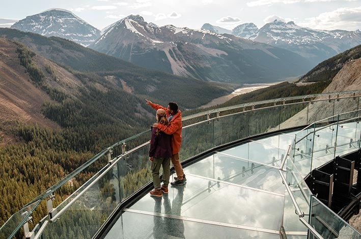 Two people look out over a wide valley from a glass platform.