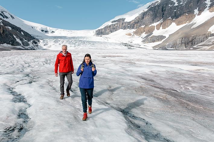 Two people walk on a glacier of ice below ice-covered mountains.