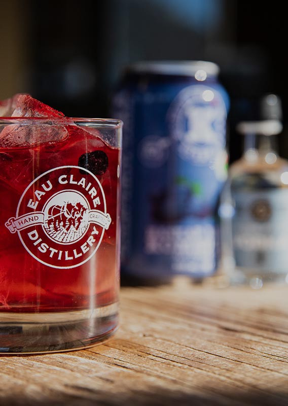 A red cocktail in a small glass on a wooden table with other beverages slightly out of focus.