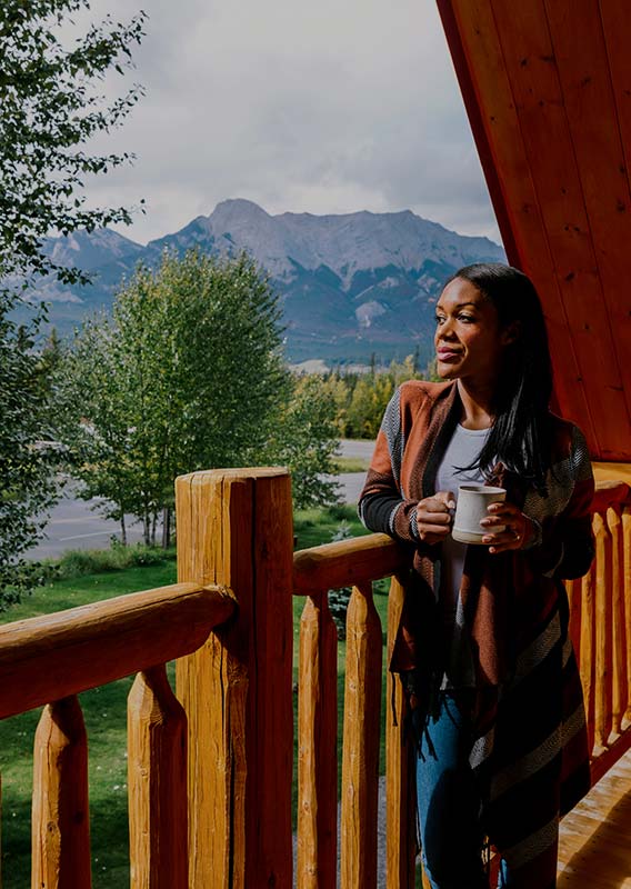 A woman stands on a wooden balcony looking out towards a forest and mountains