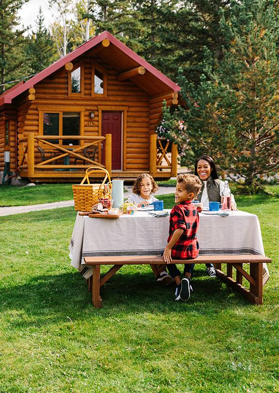 A family at a picnic table and barbecue, near wooden cabins.