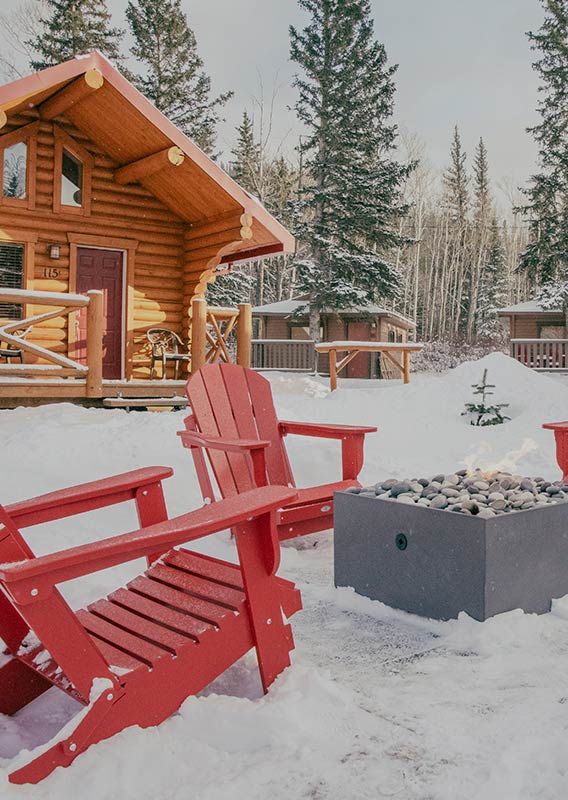 Red chairs positioned around a firepit in the winter between wooden cabins.