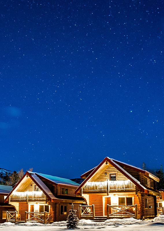 Wooden cabins with red roofs covered in snow, lit up in the night.