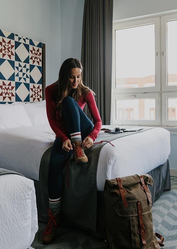 A person ties up their hiking boots on a hotel bed.