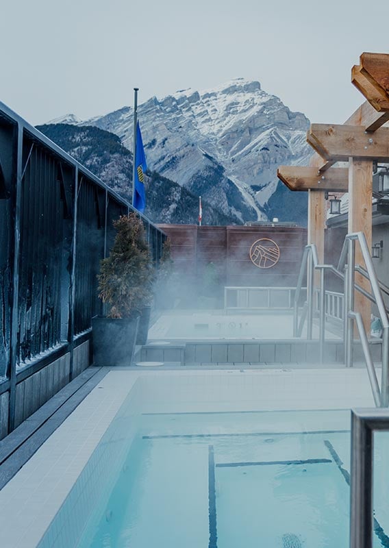 Two steaming hot tubs on a rooftop with a view towards a snow-covered mountain.