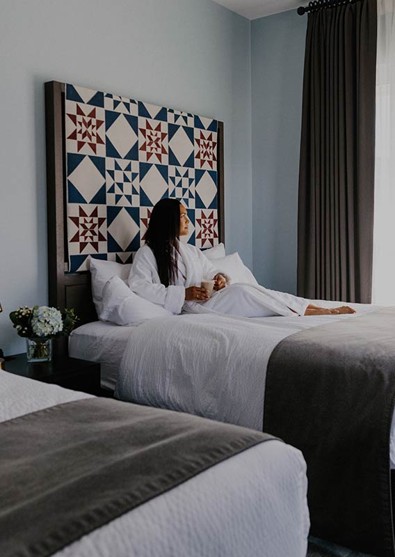 Woman sitting in a white bathrobe on her bed in a hotel room.