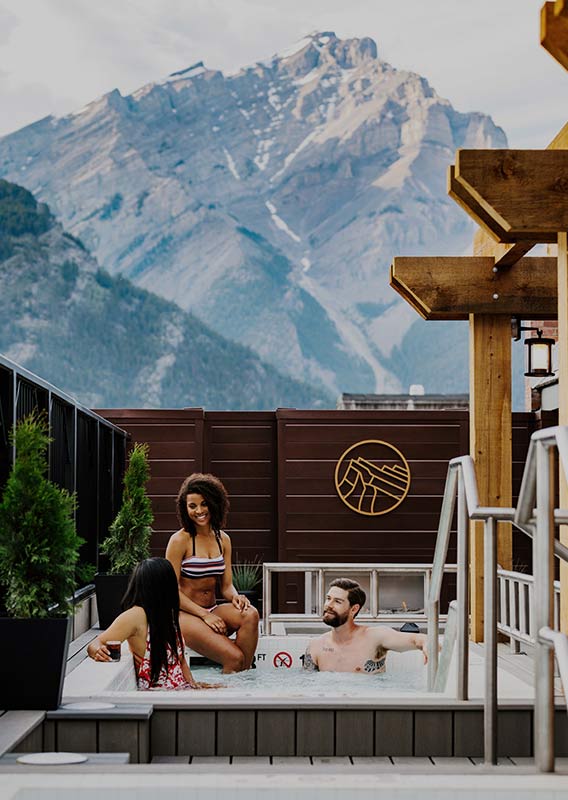 People enjoy the rooftop hot tubs at the Mount Royal Hotel with Cascade Mountain behind them
