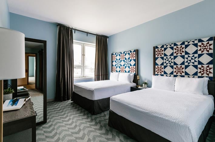 Two beds with red and blue quilted headboards, with sun shining through a window.