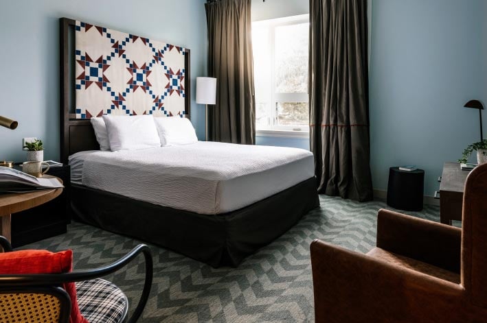 A bed with quilted headboard under a window, with chairs around.