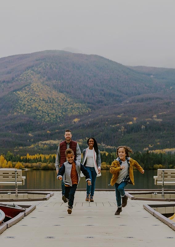 A family walking along a dock on a calm lake below tall mountains.