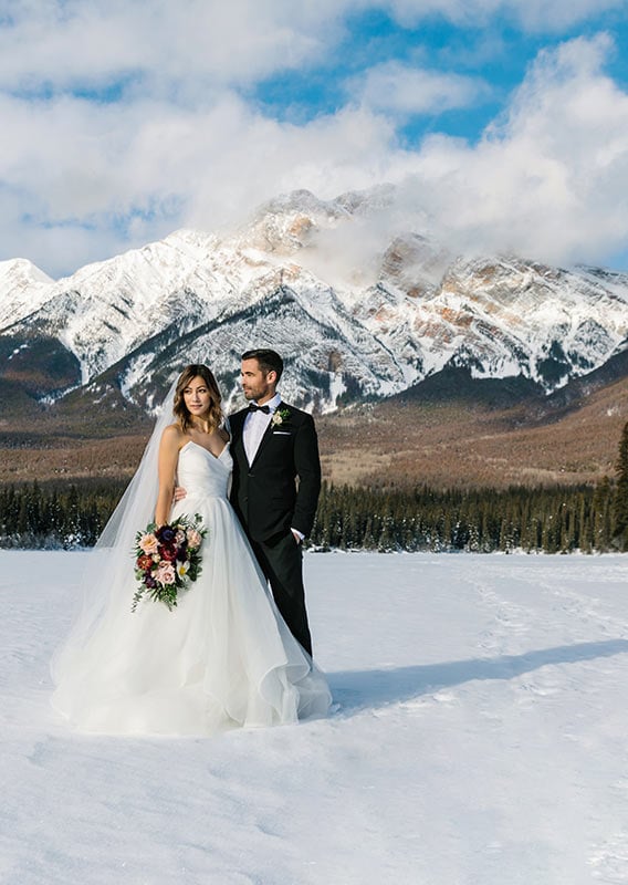 A bride and groom pose on a frozen Pyramid Lake at the base of a snowy mountain