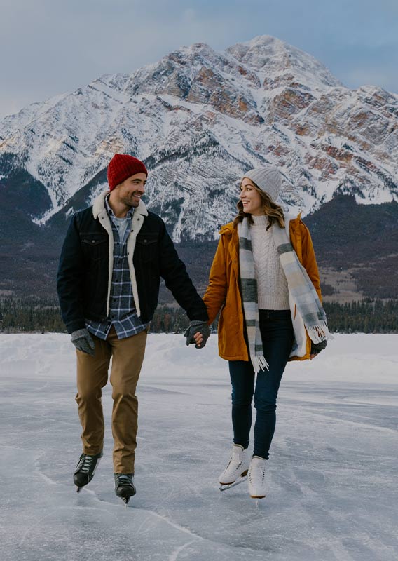 A couple holds hands while skating on a frozen lake. Snow-covered mountains surround.