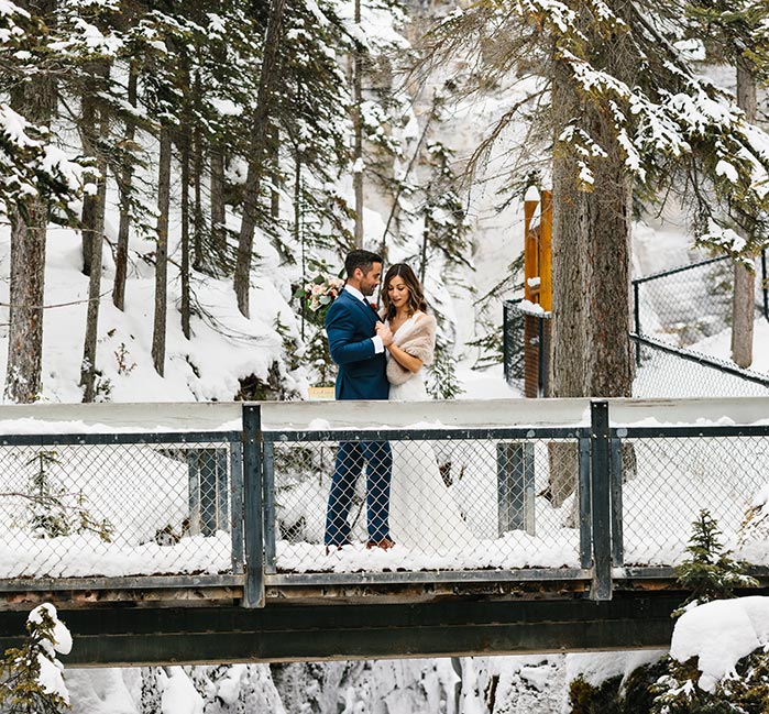 A bridge and groom stand on a snowy bridge in the forest