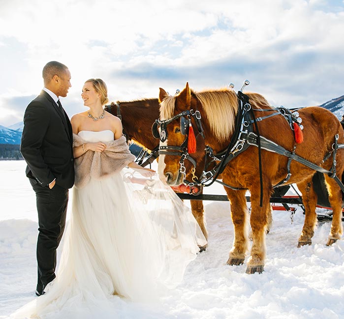 A bridge and groom stand in the snow with a horse drawn sleigh next to them