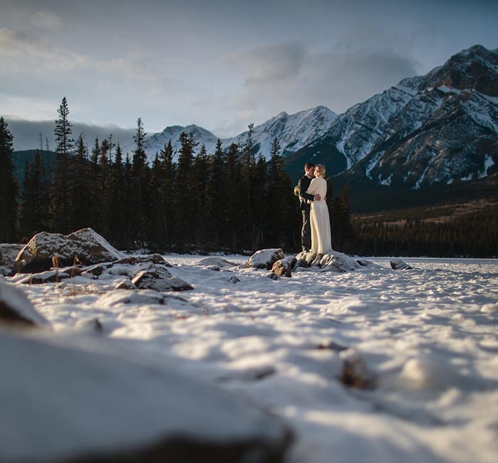 A newlywed couple stands on a frozen lake below tree-covered mountains