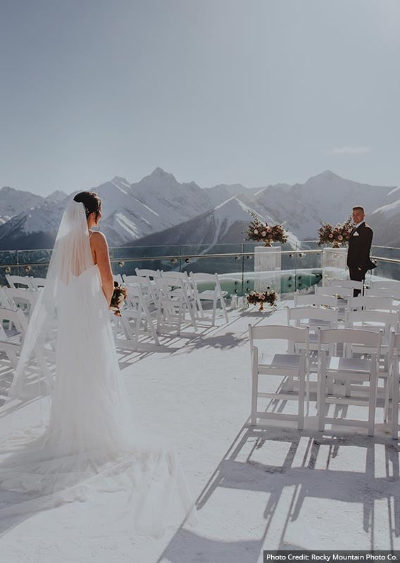 A bride walks between chairs set up for a wedding on a terrace overlooking snow-covered mountains.