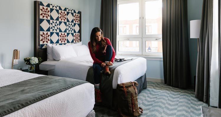 A woman ties her boot laces on a bed in a sunny hotel room.