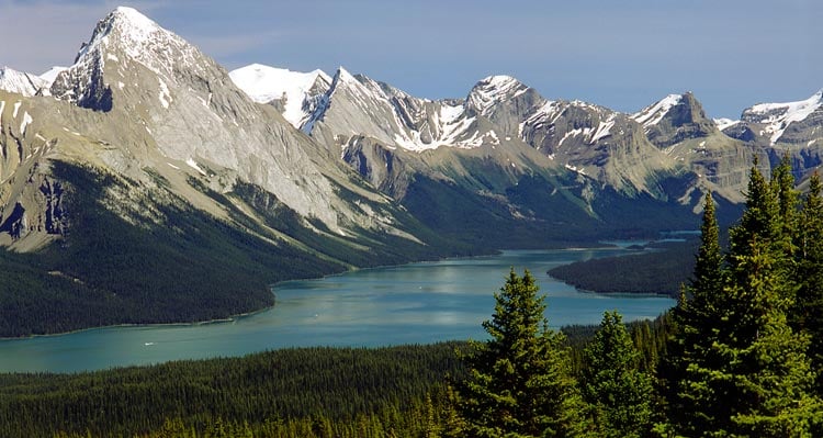 A blue lake sits below snow-capped peaks and forested mountain-sides