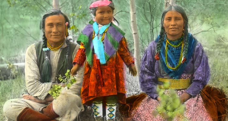 Two adults and a child wearing traditional Nakoda attire among aspen trees.