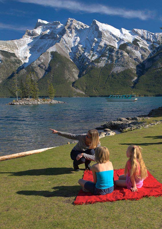 Two groups of people enjoy picnics on grass and views of mountains across a lake