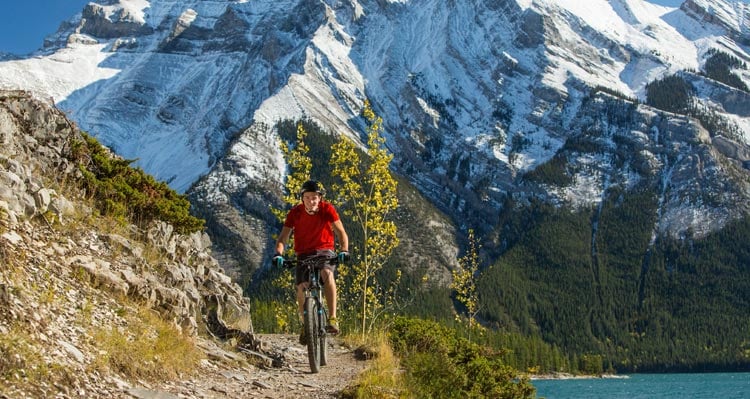 A cyclist rides on a narrow dirt trail above a blue lake with snow-covered mountains behind.