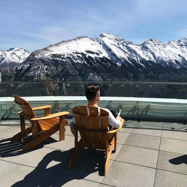 A person sits in a wooden Adirondack chair looking out towards snow-covered mountains.