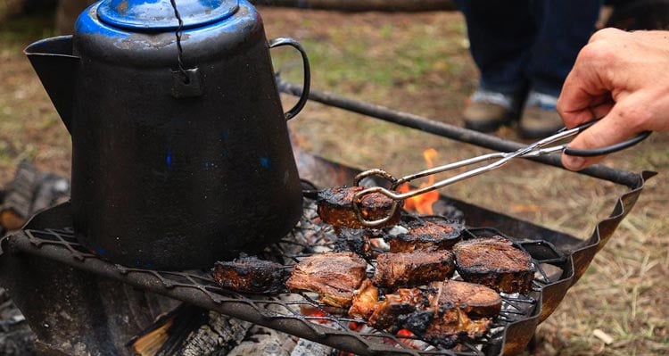 A coffee can and barbecuing meat sit on a grill over an open campfire.