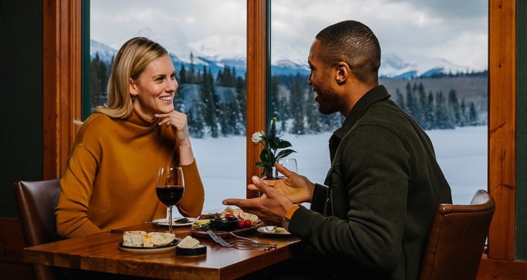 Two women share a cheers at a window table looking out to tree-covered mountains.