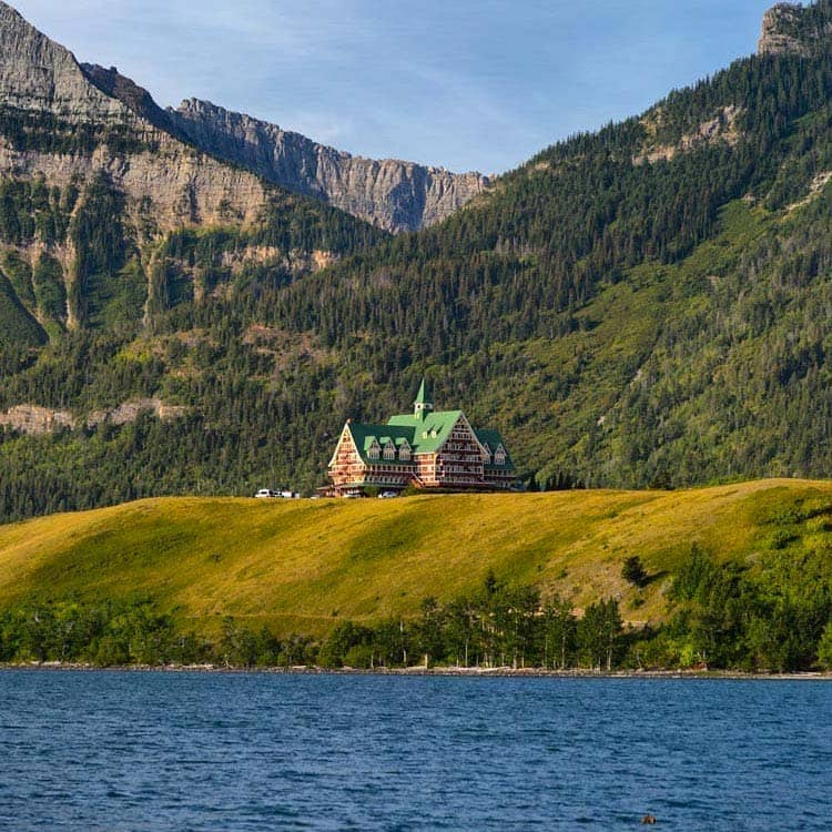 The Prince of Wales Hotel sits atop a green hill below forest-covered mountains.