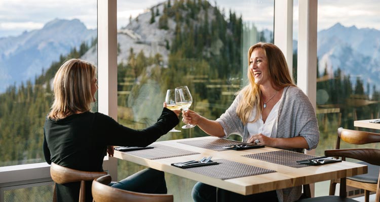 Two women share a cheers at a window table looking out to tree-covered mountains.