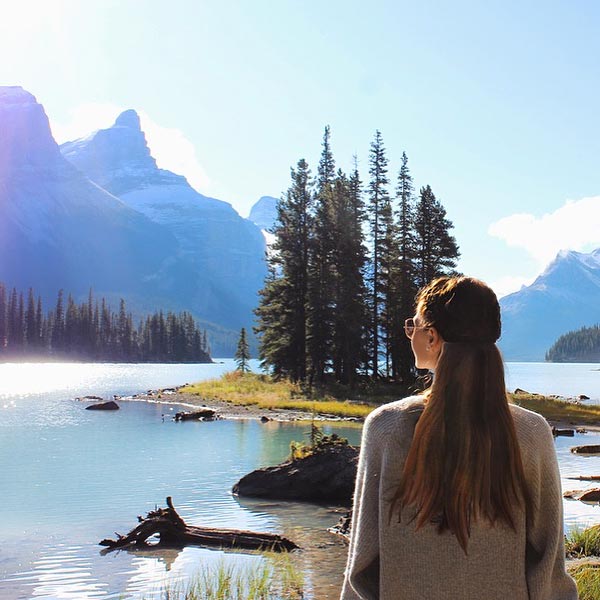 A person stands at a viewpoint looking towards a turquoise mountain lake and a small pine tree-covered island