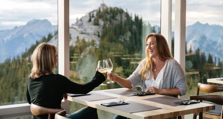 Two women raise wine glasses in a cheers at a window seat, looking out towards tree-covered mountains.