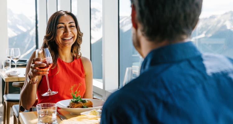 Two people dine at a wide window overlooking mountains.