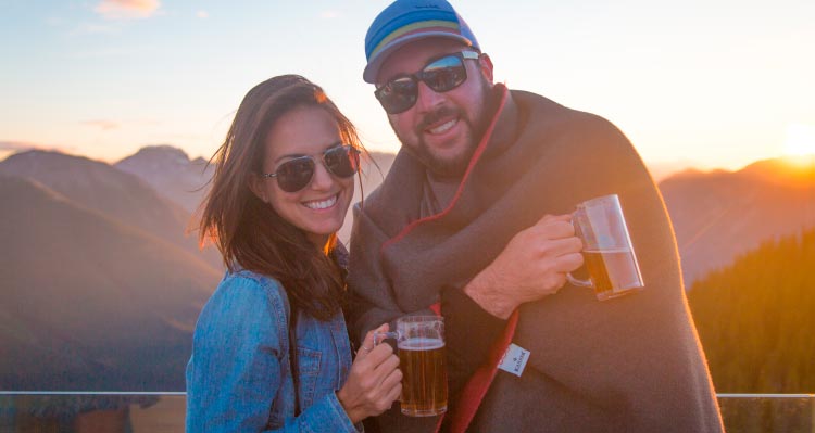 A pair of people stand in the glowing light of the sunset wrapped in a blanket.