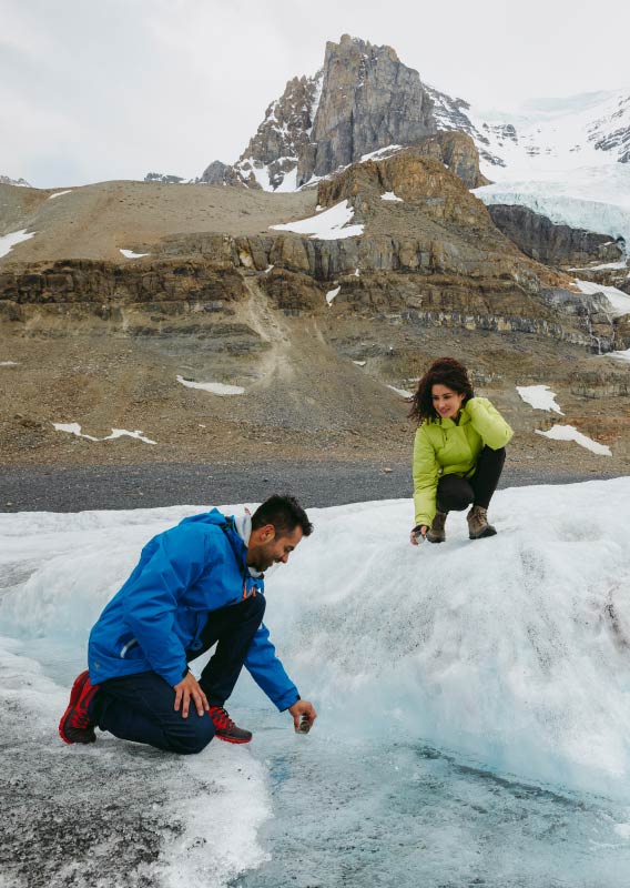 Two people crouch on a glacier, looking at and touching flowing glacial water.