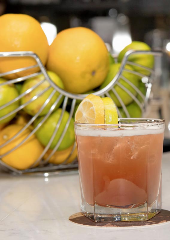 A pink cocktail sits on a table in front of a bowl of lemons and limes.