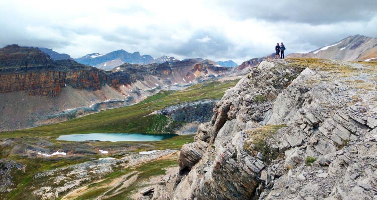 Two people stand atop a high cliffside above a green alpine meadow and small blue lake