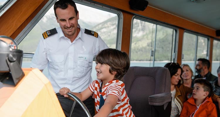 A child sits in the captain's chair of a boat with the boat captain.