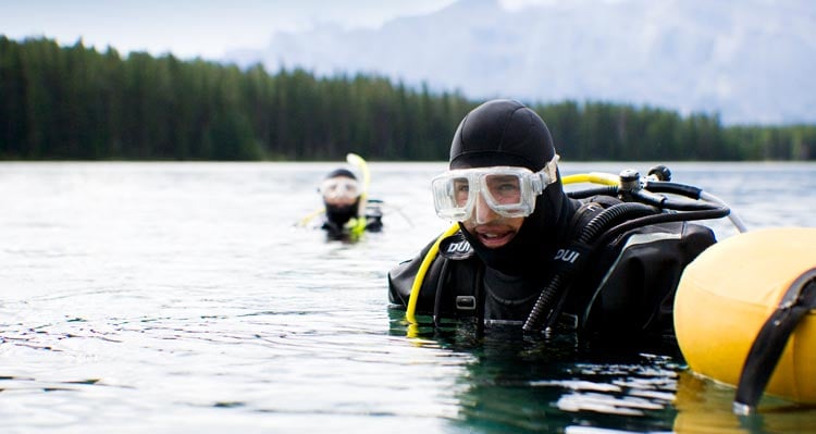 Two SCUBA divers prepare to dive on a lake surrounded by forests and mountains.