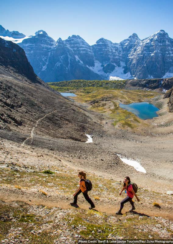 Two hikers walk along a trail above a blue lake and wide valley