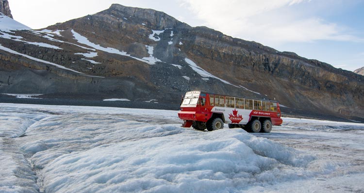 A red and white Ice Explorer bus sits on a field of ice with a mountain rising behind.