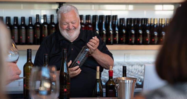 A bearded man smiles holding a bottle of wine at a wine bar.