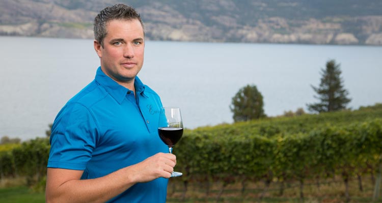 A man in a blue shirt holds a glass of wine in front of a lake.