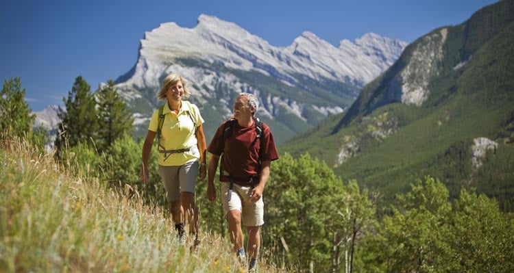 Two people hike along a hillside meadow with mountains all around.