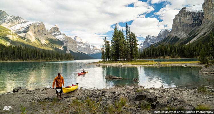 Two kayakers approach the shore near a tree-covered peninsula.