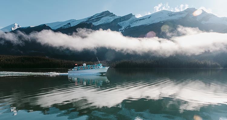 A boat cruises along a crisp lake below a low cloud and snow-covered mountains.